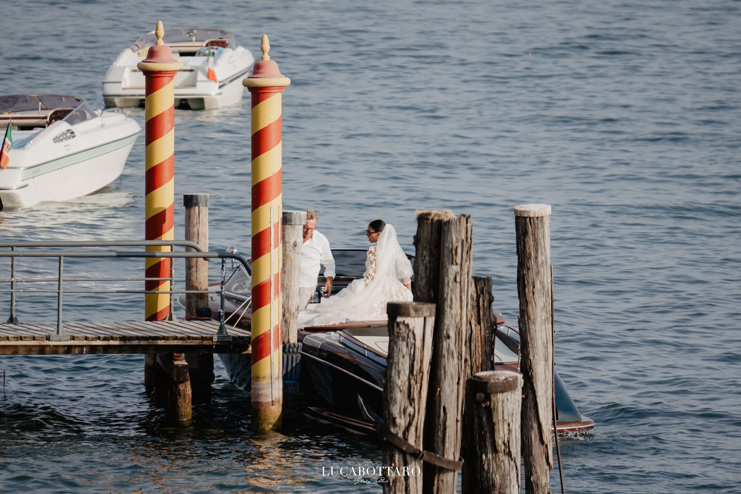 Sposa con il velo seduta su un motoscafo d'epoca ormeggiato al pontile, tra i pali veneziani rossi e gialli