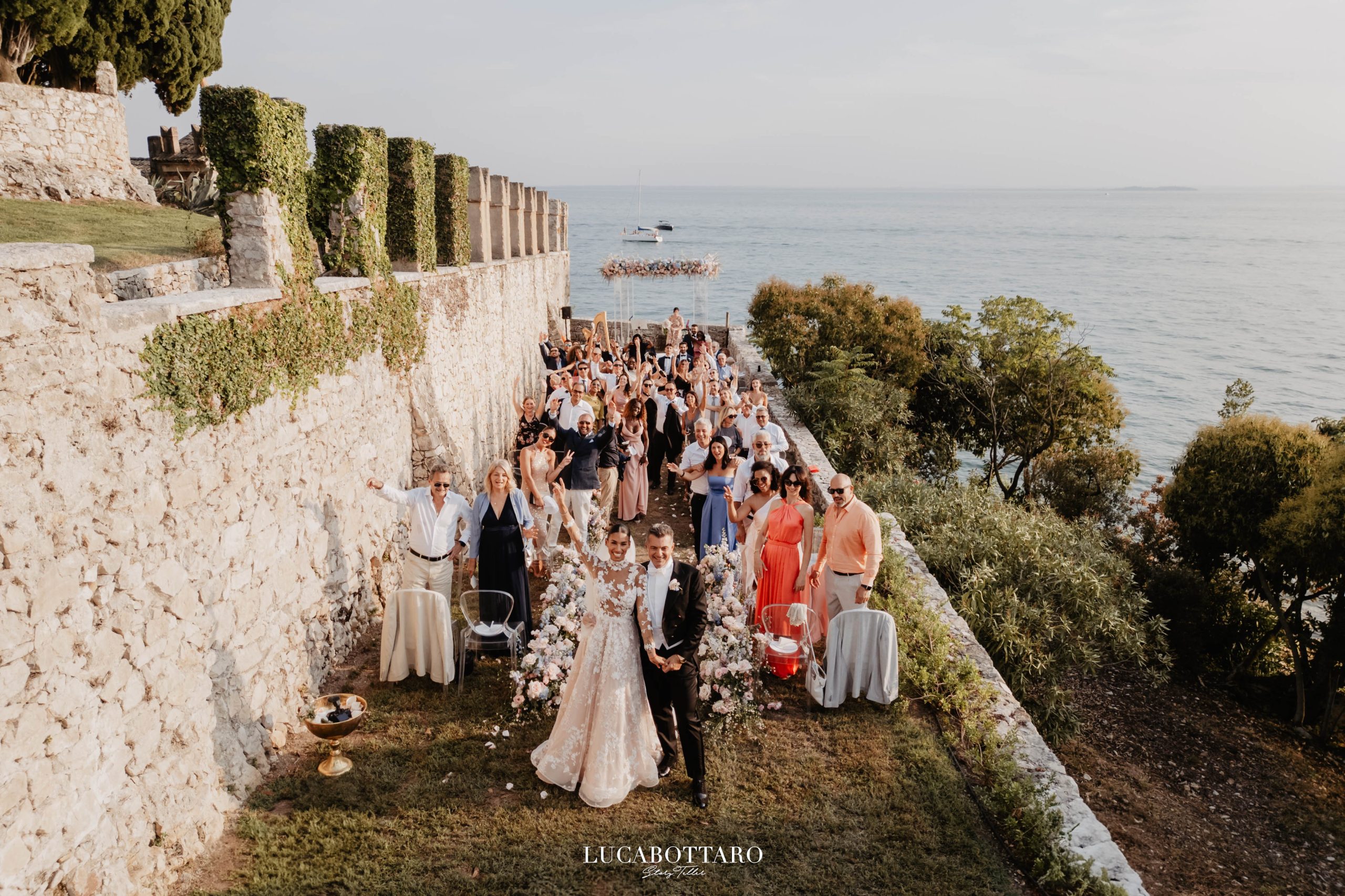 Veduta dall'alto della cerimonia a Villa San Vigilio: sposi in fondo alla navata, invitati in festa, mura merlate e lago