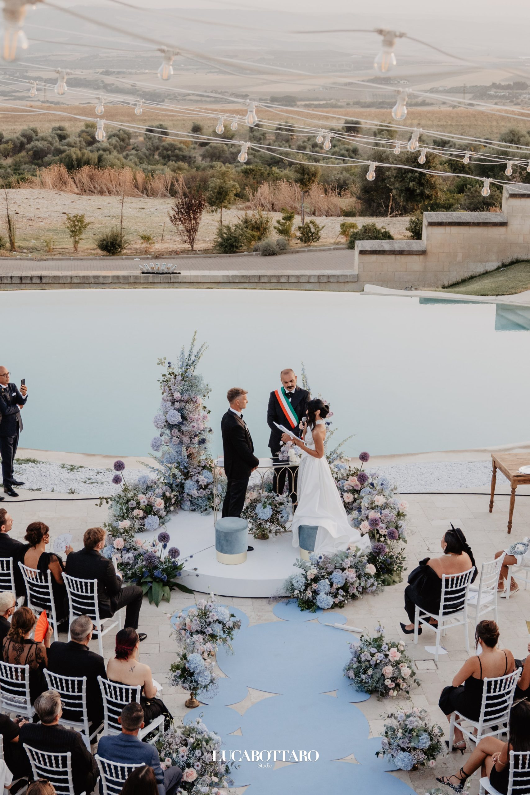 Civil wedding ceremony from above, with the couple facing the officiant on a floral circle beside an infinity pool and olive hills